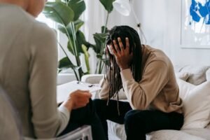 Anxious African American male with hands on head sitting on sofa near crop female psychologist with clipboard during appointment in office